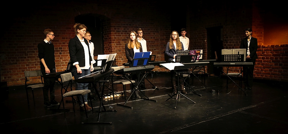 Zdjęcie przedstawia ośmiu muzyków stojących na scenie przy swoich instrumentach. 