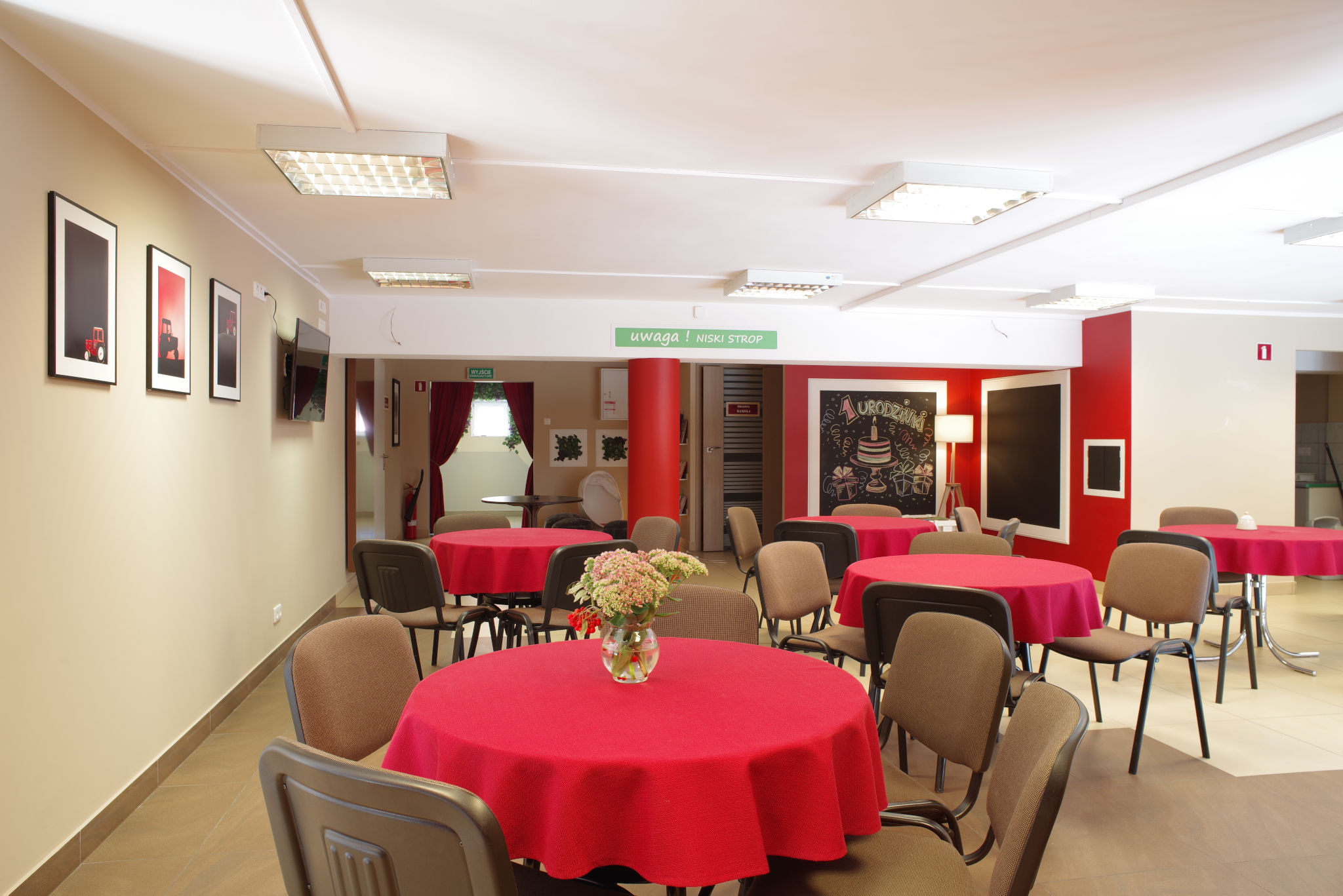 Sala na spotkanie, na pierwszym planie kilka stolików kawiarnianych, okrągłych z czerwonym obrusem. Rozstawione krzesła wokół. Na ścianie obrazy w antyramach.