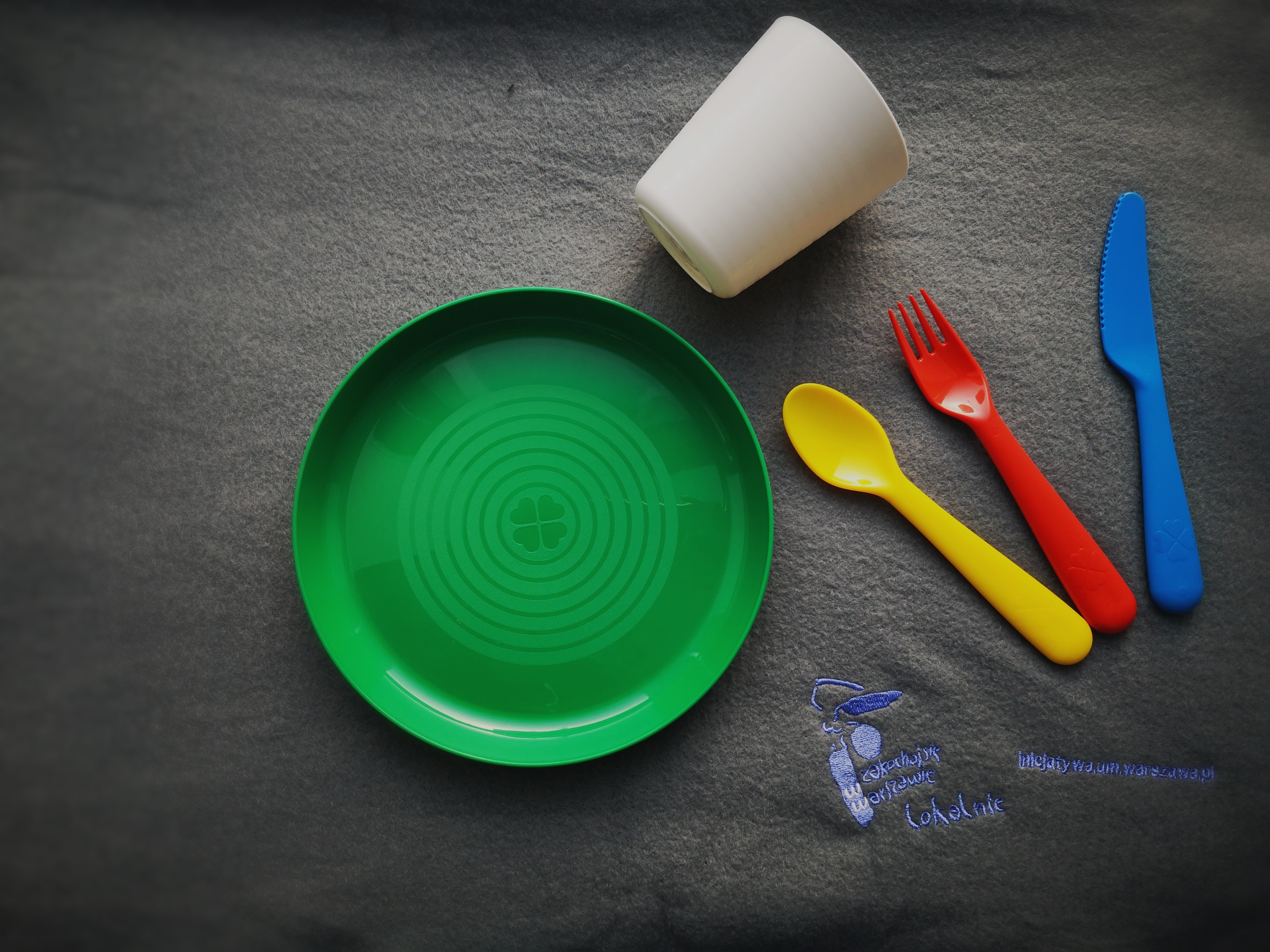 Plastikowy zestaw naczyń: talerzyk, kubek, zestaw sztućców. Każdy z przedmiotów jest w innym kolorze.